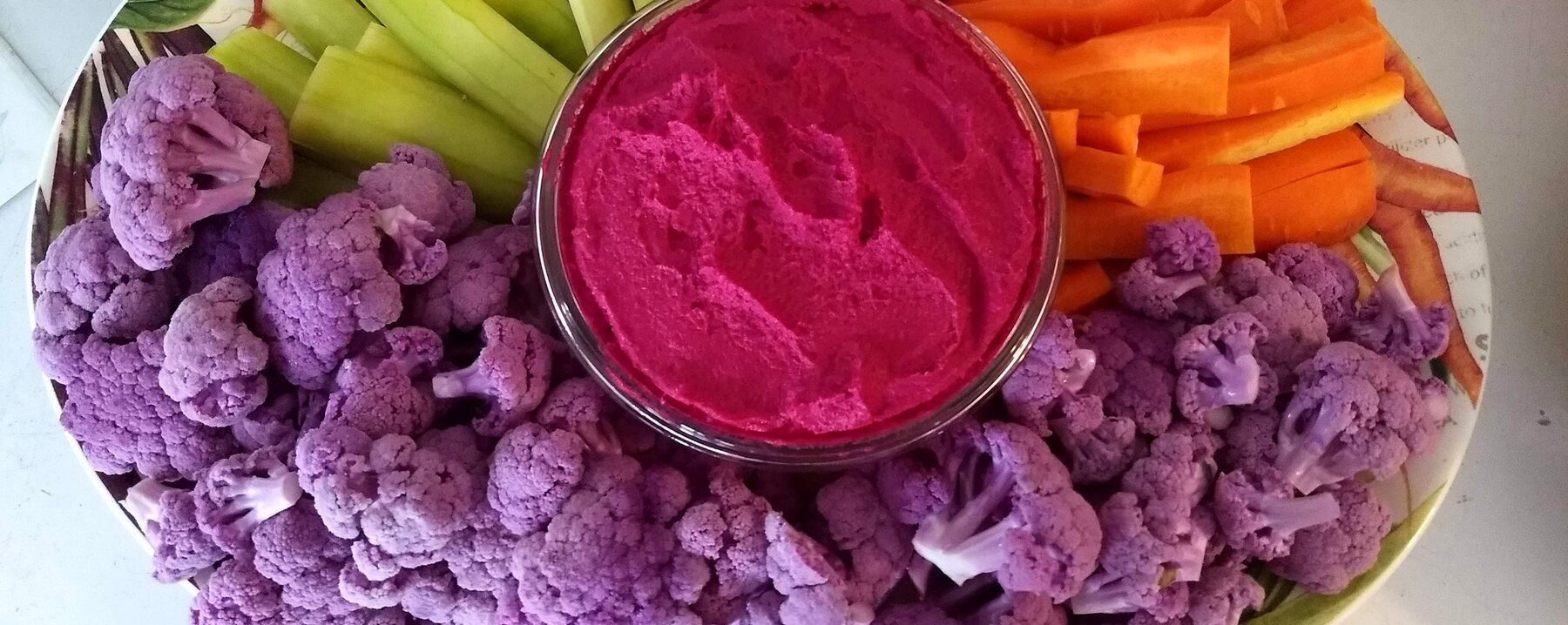 Beet hummus surrounded by purple cauliflower, carrots, and cucumber